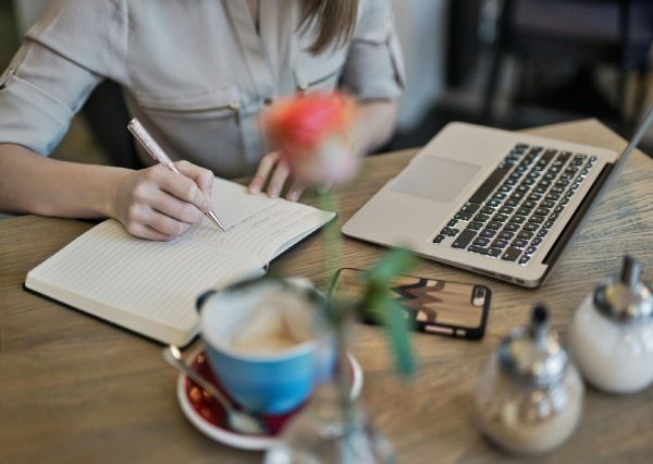 A person writes on a notebook beside a MacBook.